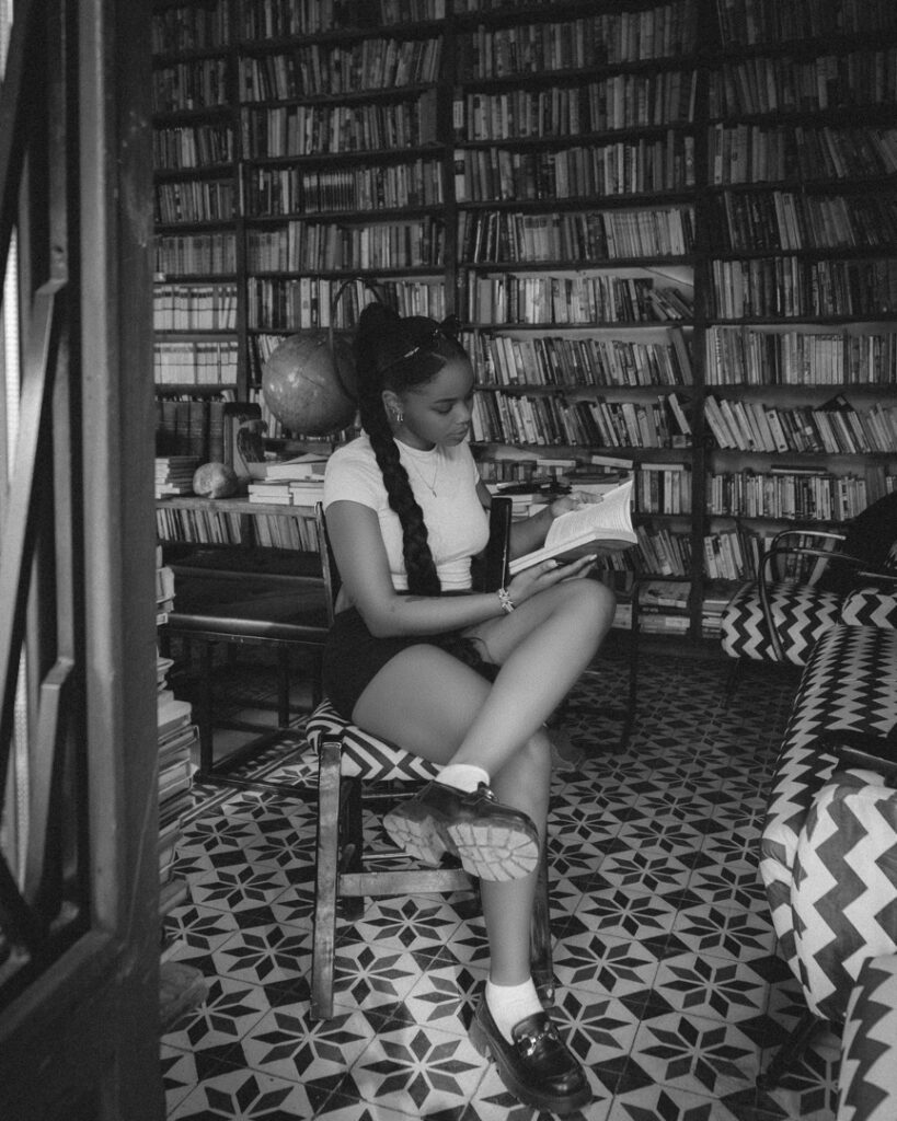 Eine junge Schwarze Frau sitzt gemütlich in einer Bibliothek, Bücherregal im Hintergrund.